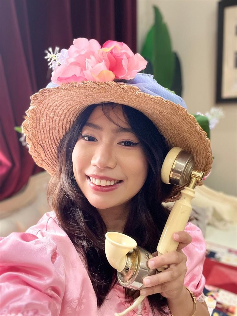 Thiri holding an antique candlestick telephone, wearing a straw hat with pink roses