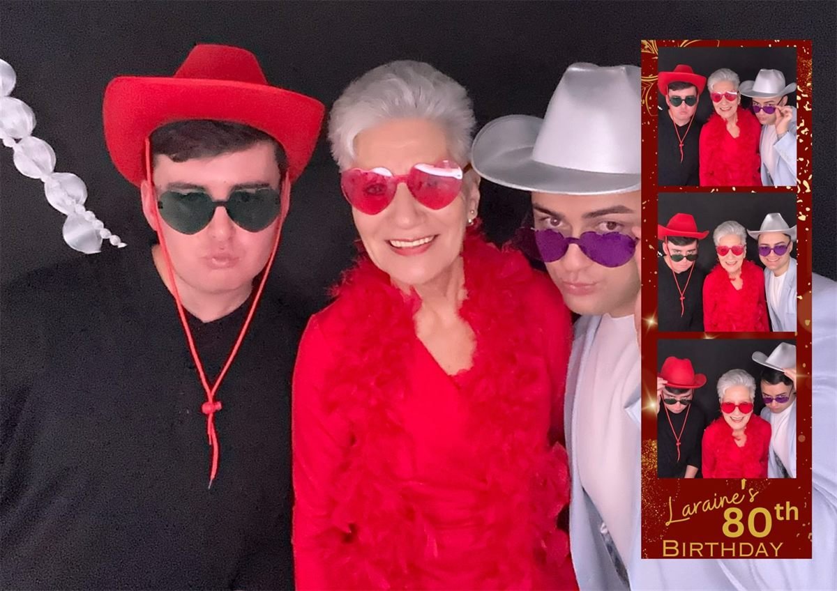 Three generations posing in feather boas, hats and heart-shaped sunglasses against a black backdrop at an 80th birthday in Malaga, with their red and gold photo strip alongside